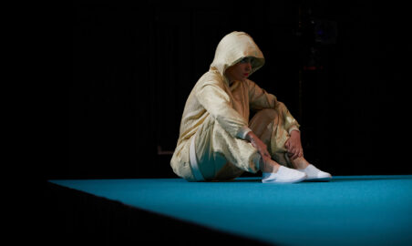 Against a dark background, a person in a pae, loose-fitting hooded tracksuit sits up, knees bent to their chest, on a section of blue floor.